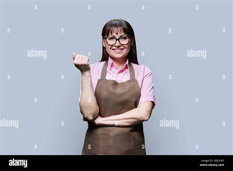 Positive Smiling Mature Woman In Apron On Gray Studio Background Stock