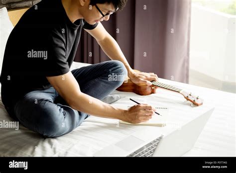 Happy Young Asian Man Playing Ukulele Sitting On Bed In Bedroom Stock