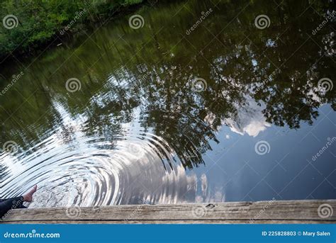 Feet Dangle In Woodland Pond Water From A Wooden Dock Making Circlular