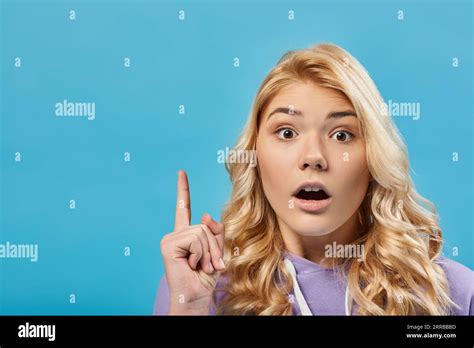 Portrait Of Amazed Blonde Teenage Girl With Open Mouth Showing Idea Gesture On Blue Stock Photo