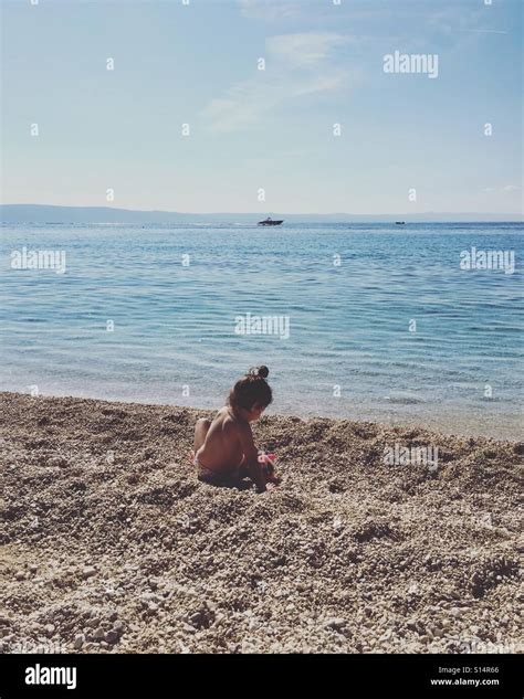 Cute Little Girl Playing On The Pebble Beach Close To The Calm Blue Sea