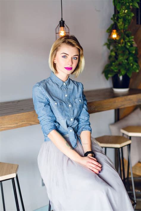 Shy Sweet Girl With Blonde Hair And Pink Lips Sitting In A Coffee Shop With Wooden Chairs And