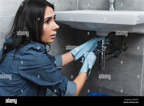 Worried Brunette Girl In Denim Shirt Repairing Sink Pipe With Wrench