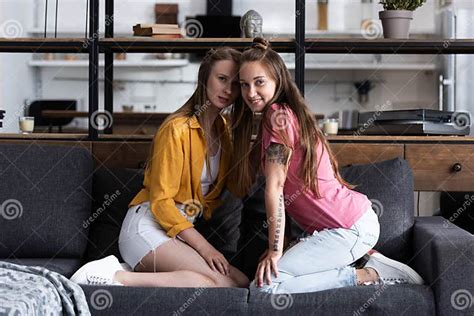 Two Smiling Lesbians Sitting On Sofa And Looking At Camera In Living