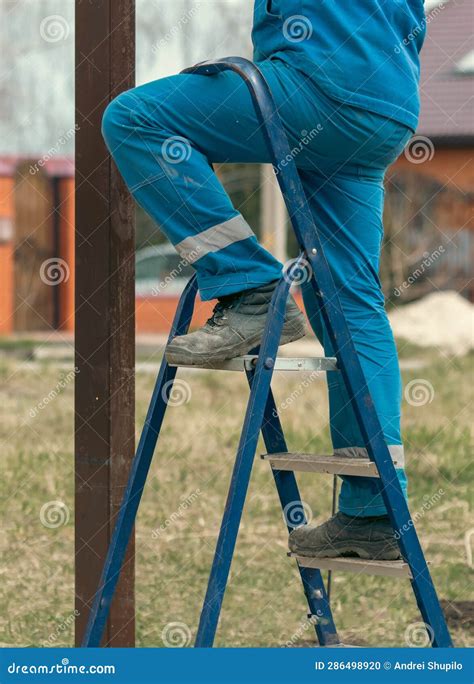 Worker In Blue Overalls Stands On A Metal Ladder In The Yard Stock