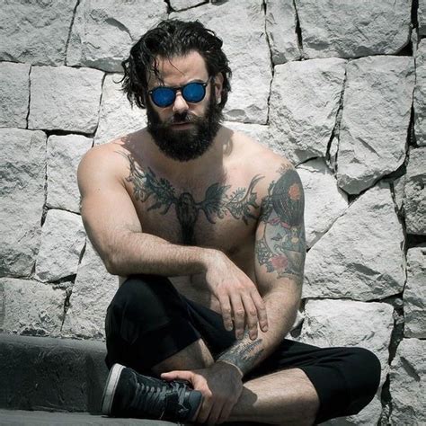 A Shirtless Man With Tattoos And Sunglasses Sitting On A Stone Wall Looking At The Camera