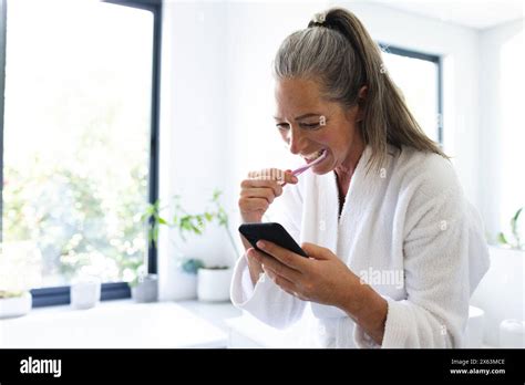 At Home Mature Caucasian Woman Brushing Her Teeth Checking Smartphone