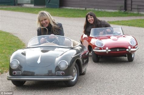 Two Women Are Sitting In Toy Cars On The Road One Is Red And The Other