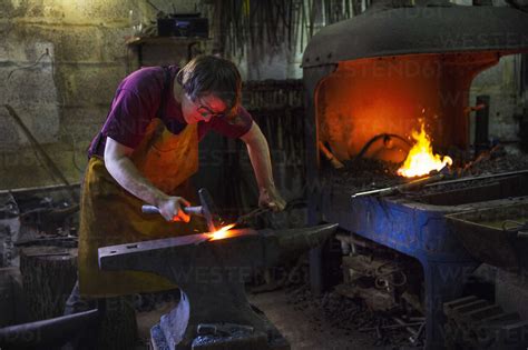 A Blacksmith Striking Red Hot Metal On An Anvil Inside A Workshop Stock Photo