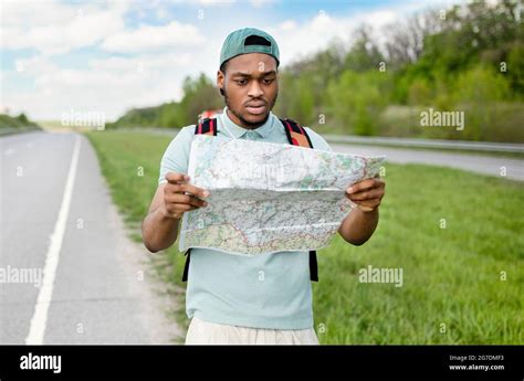 Unhappy young black man hitchhiking on road, looking at map, feeling ...