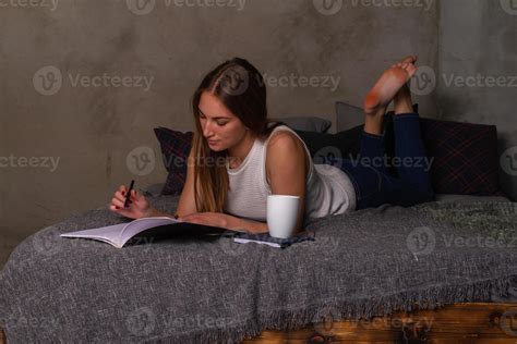 Woman Lying On Her Stomach In Bed And Writing Notes In Notebook