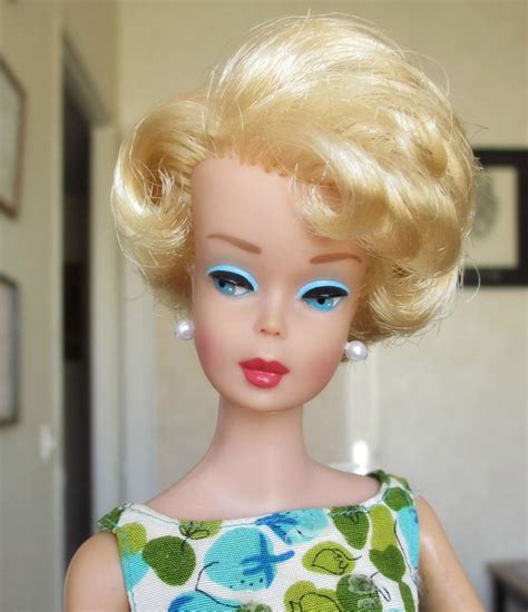 A Doll With Blonde Hair And Blue Eyes Is Wearing A Green Floral Dress In Front Of A White Door