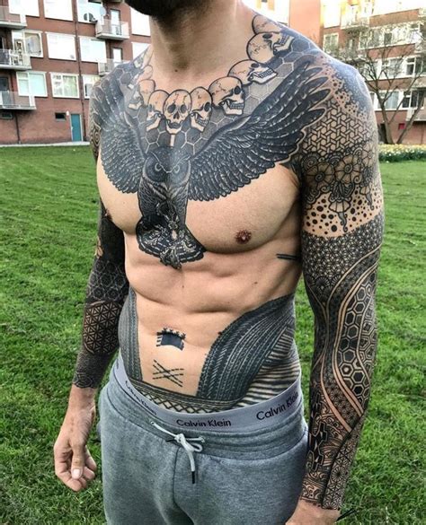 A Man With An Eagle Tattoo On His Chest Standing In Front Of Some Grass And Buildings