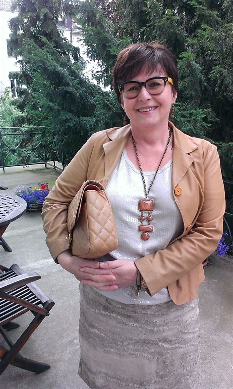 A Woman Wearing Glasses And A Tan Jacket Is Holding A Purse In Front Of Her