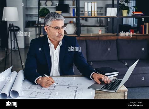 Mature Architect Working With Laptop And Drawings In Office Stock Photo