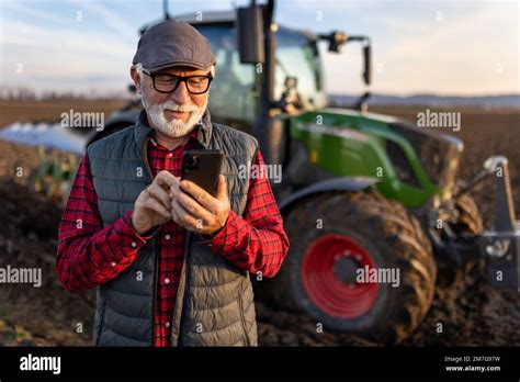Smiling Mature Farmer Looking At Mobile Phone In Front Of Tractor In