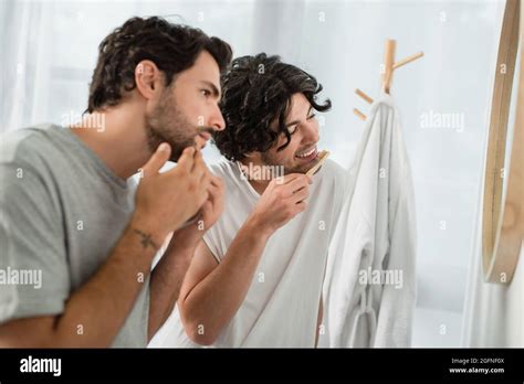 Blurred And Tattooed Gay Man Touching Face While Boyfriend Brushing Teeth And Smiling In