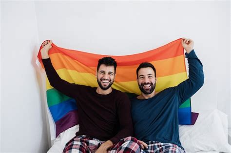 Casal gay masculino segurando a bandeira do arco íris lgbt dentro de casa na cama em casa
