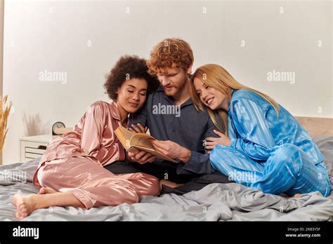 Open Relationship Concept Redhead Man Reading Book Next To Happy Multicultural Women In Pajamas