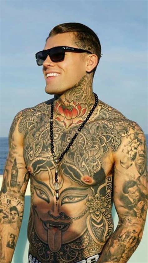 A Man With Tattoos On His Chest Standing Next To The Ocean Wearing Sunglasses And Beads