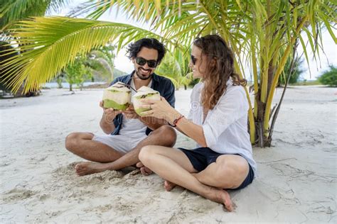 A Beautiful Young Couple Joyfully Sits On White Sand Under A Palm Tree