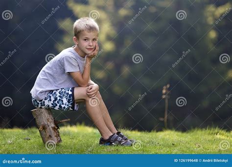 Young Cute Blond Child Boy Sitting On Tree Stump On Green Grassy Stock