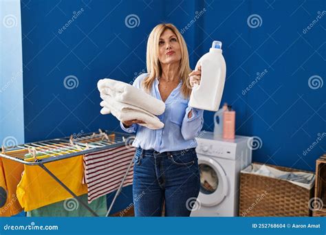 Middle Age Blonde Woman Holding Laundry Basket And Detergent Bottle