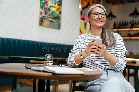 Mature Grey Woman Smiling And Using Cellphone While Sitting In Cafe