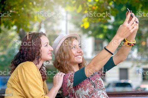 Two Women Taking Selfies While Sitting On A Park Bench Stock Photo