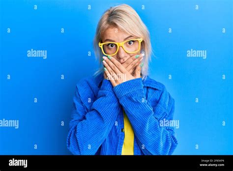 Jeune fille blonde portant des vêtements décontractés et des lunettes choqué couvrant la bouche