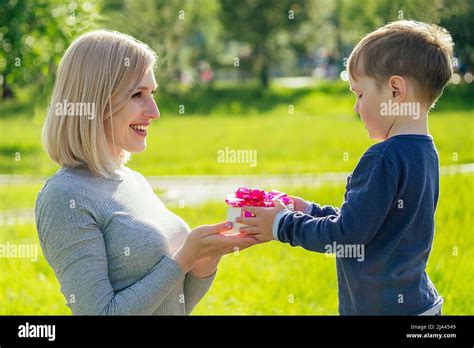 Cute Baby Son Gives A Gift Box To His Beautiful Blonde Mother In The Park On A Background Of