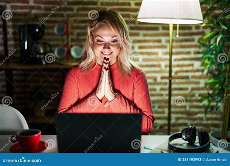 Blonde Woman Using Laptop At Night At Home Praying With Hands Together