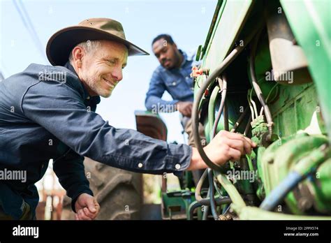 Smiling Mature Farmer Repairing Engine While Colleague Sitting On