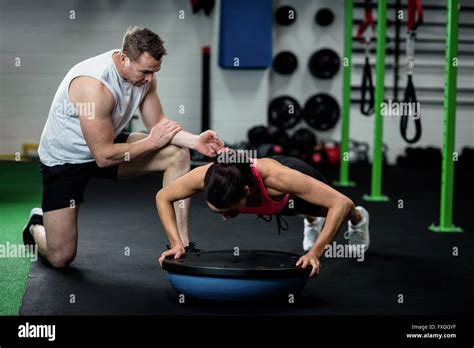 Trainer Assisting A Woman Doing Push Ups On A Bosu Ball At Gym Stock