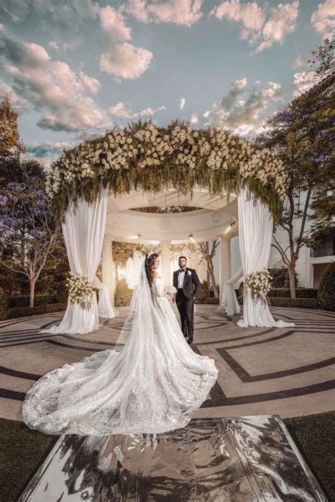 A Bride And Groom Standing In Front Of An Arch With Flowers On It At