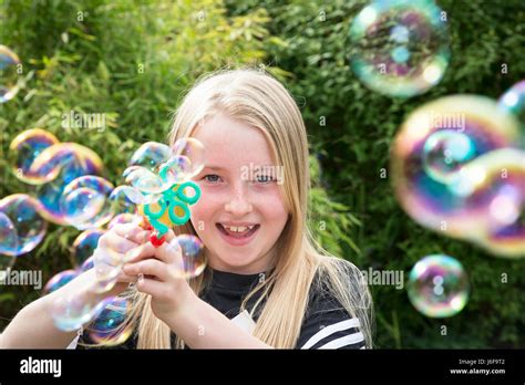 Ten Year Old Blonde Girl Uses A Small Machine To Blow Bubbles At The Camera In A Garden Stock