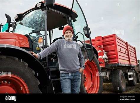 Smiling Mature Farmer Standing With Hand In Pocket Against Red Tractor