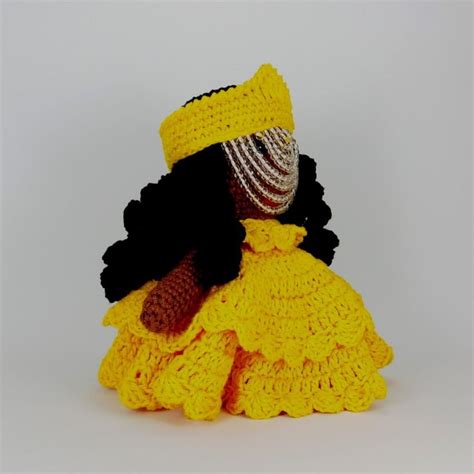 A Crocheted Doll Is Dressed In A Yellow Dress And Black Hat Sitting On