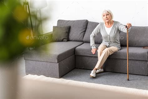 Senior Woman With Walking Stick Sitting On Sofa In Living Room Stock