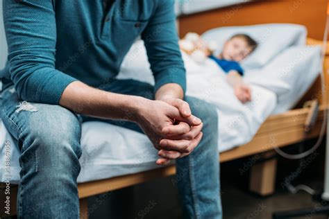 Partial View Of Dad Sitting Near Sick Son In Hospital Bed Stock Photo