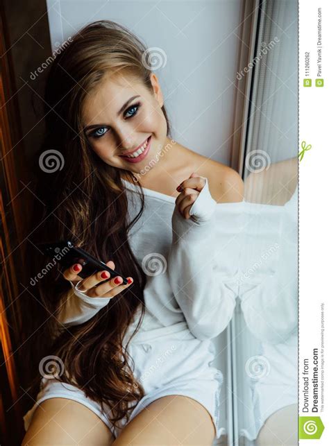 Pretty Brunette Girl Sitting On Window Sill With Smartphone In Hands Long Wavy Hair Stock Photo
