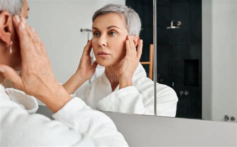 Mature Woman In Bathrobe Looking Herself In The Bathroom Mirror Stock