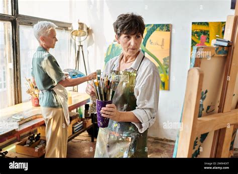 Talented Mature Woman With Paintbrushes Looking At Easel Near Female
