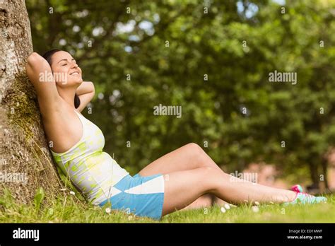 Smiling Fit Brunette Sitting And Relaxing Against A Tree Stock Photo