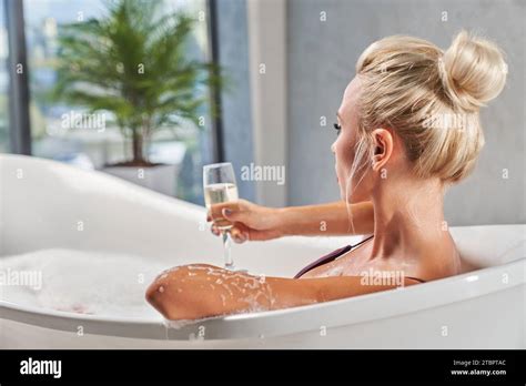 Unrecognizable Blonde Woman Taking Glass Of Sparkling Wine While Lying In Bathtub Back View Of