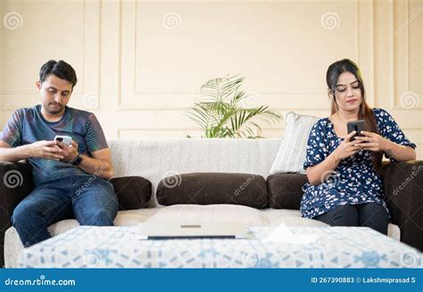 Young Indian Couple Busy Using Mobile Phone While Sitting At Sofa In