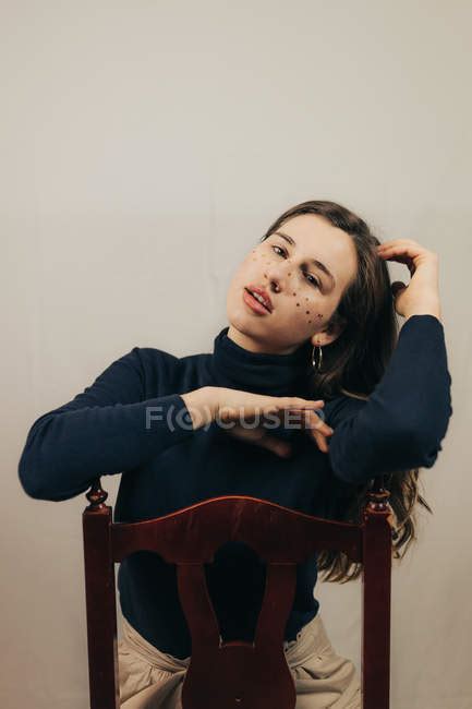 Brunette Woman With Glitters On Face Sitting On Chair And Looking At Camera Alone Indoors
