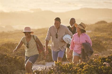 Mature Couple With Friends On Summer Vacation Walking Through Dunes On Way To Beach Carrying
