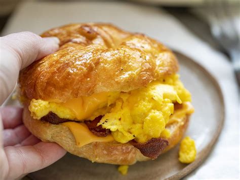 Hand Picking Up A Bacon Egg And Cheese Croissant Sandwich From Plate Breakfast Puff Pastry