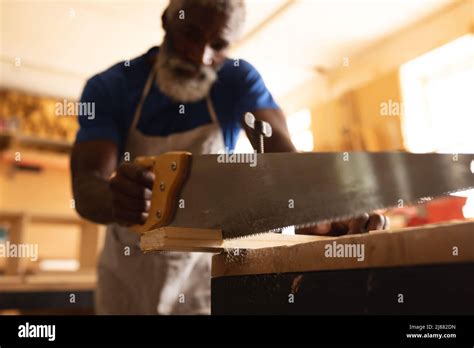 African American Mature Carpenter Cutting Plank With Saw At Workbench